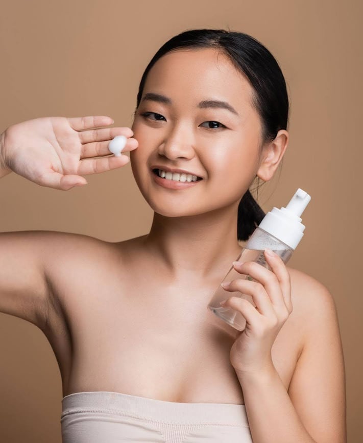 Smiling woman holds face cleanser bottle and shows foam on hand. 